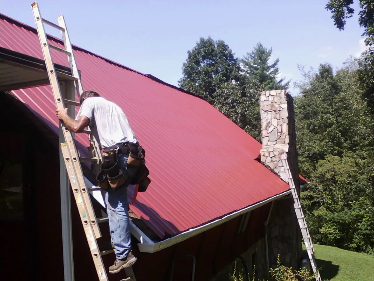 Roofer on a ladder beside a red metal roof and stone chimney — Soffit & Fascia Repair by Roofing Brothers in Ville Platte